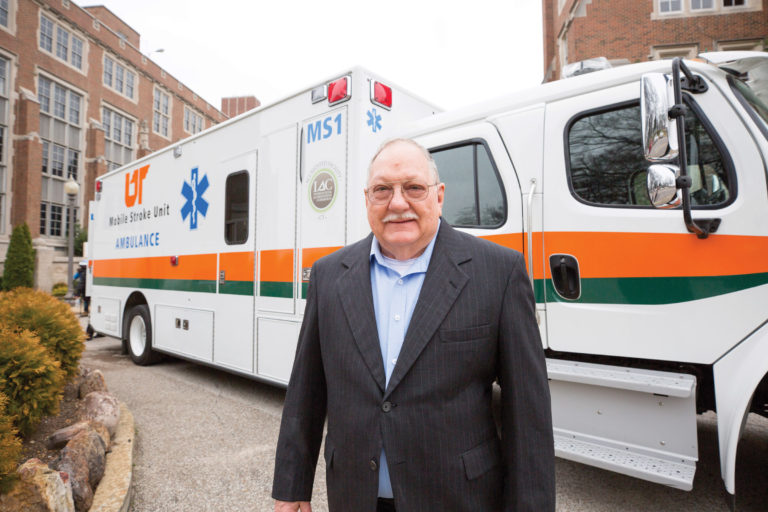 A man in business attire stands in front of a large ambulance outside a UTHSC facility