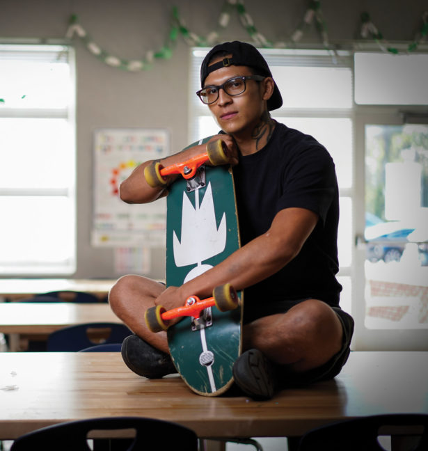 A young man sits on top of a desk holding a skateboard 