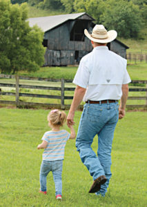 A toddler holds her father's hand as they walk the grounds of a farm.