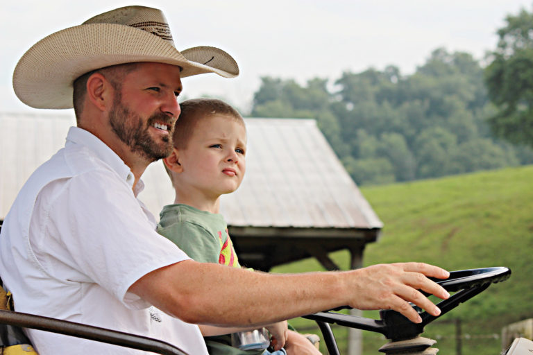 A man in a cowboy hat rides with his son is his lap on a tractor