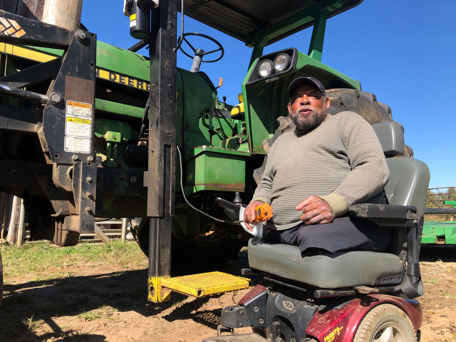 A senior Black man with an electric wheelchair next to his John Deere tractor