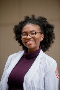 Jasmine Jefferson smiles in her burgundy turtleneck, white coat and glasses. Her curly hair is tied back