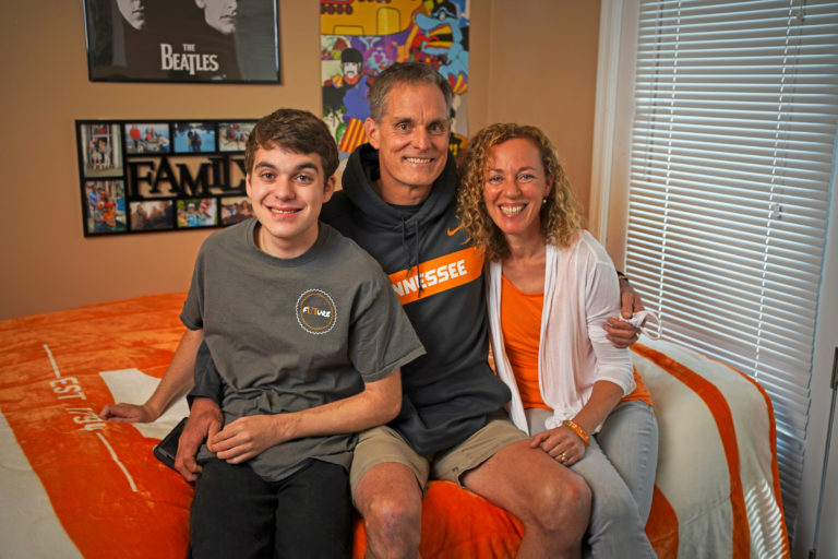 A family of three seated in their college student son's apartment. Everyone is dressed in orange, white and gray