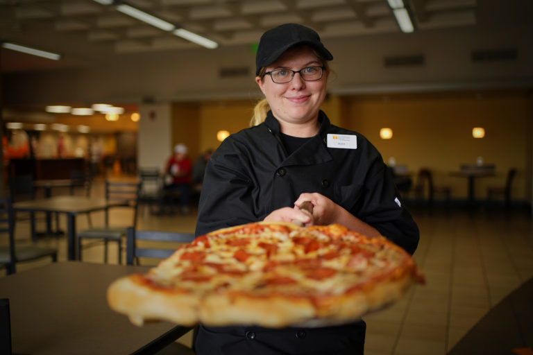 A woman in dining services uniform holds a pepperoni pizza on a tray in both bands