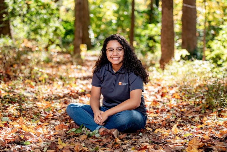 A latina woman in a gray UT polo shirt is seated on the forest floor, surrounded by fall leaves