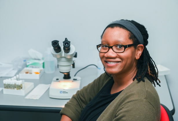 A Black woman seated in front of a microscope smiles confidently in her research lab