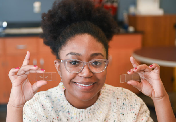 A young Black woman in an oatmeal sweater holds up miscroscope slides with bee specimens