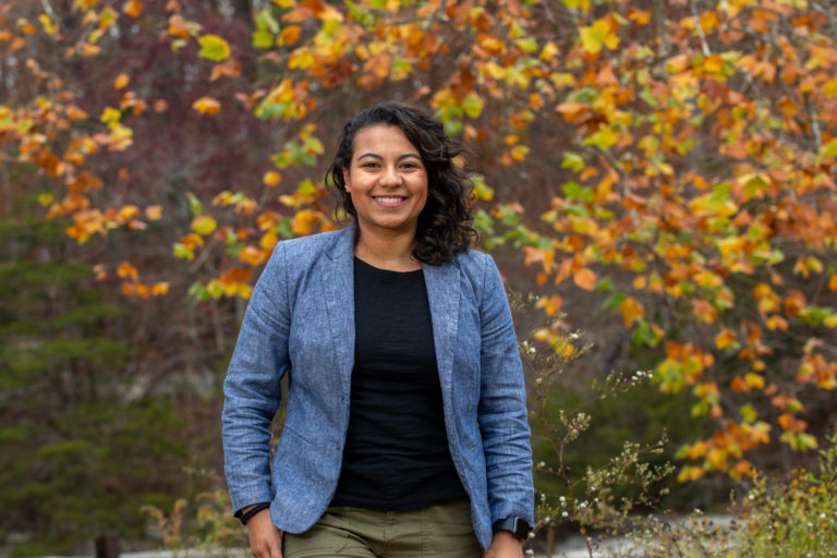A latina woman wearing a blue blazer stands in front of fall foliage